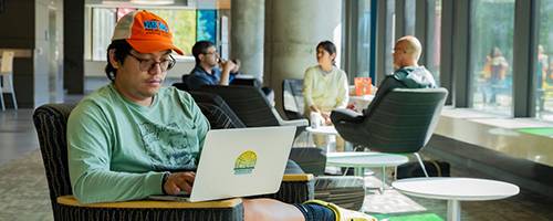 A student sits comfortably in a modern lounge area, working on a laptop. The individual is wearing a green shirt and an orange cap, with yellow Crocs on their feet.