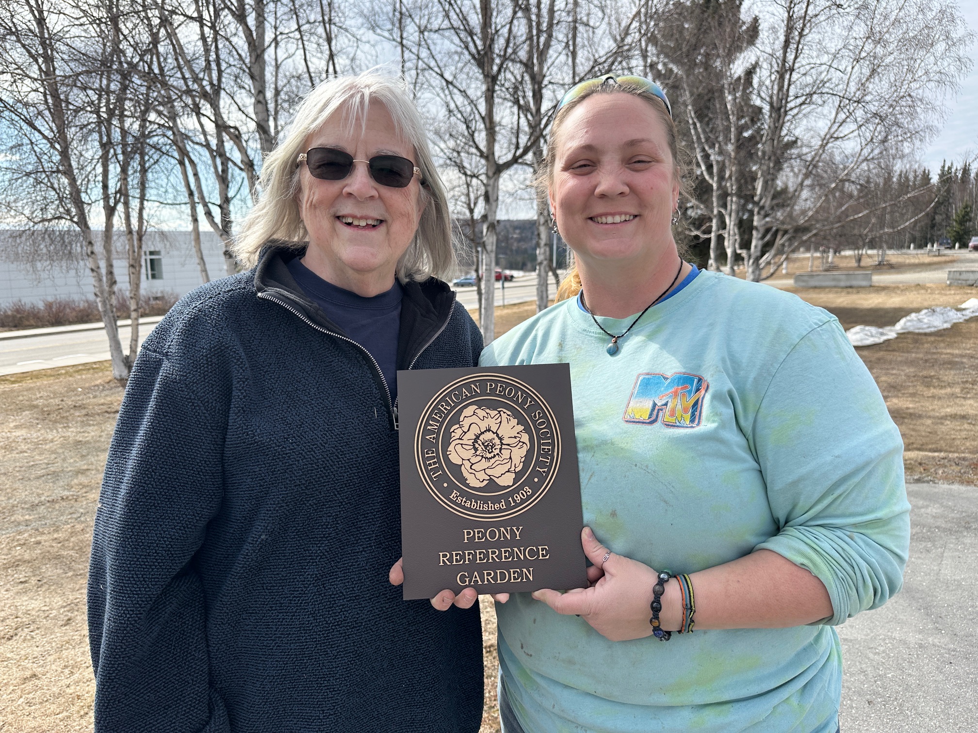 Two women stand outdoors holding a plaque that reads peony reference garden