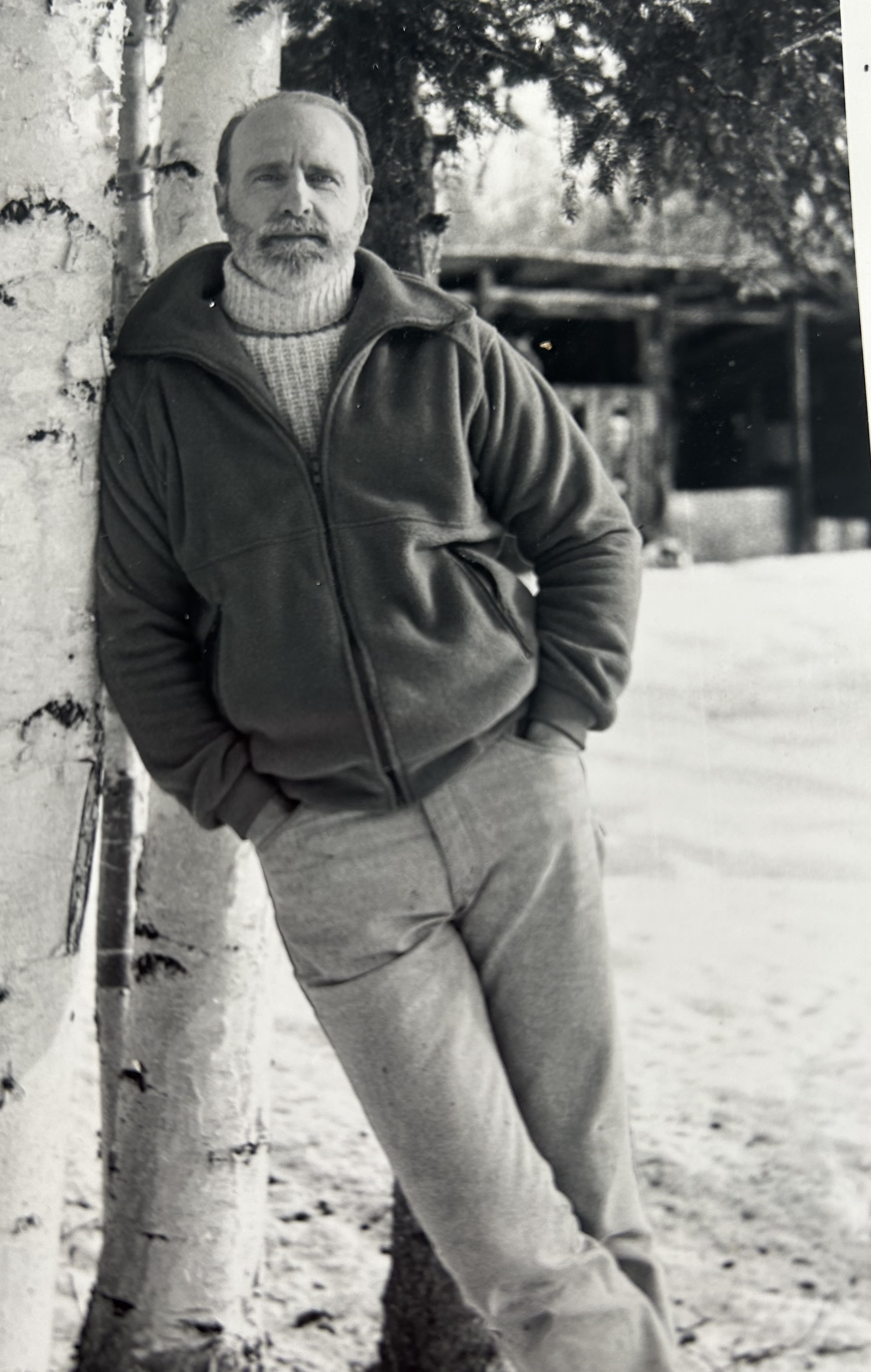 Black and white photo of Dale Guthrie in a heavy turtleneck sweater and jacket leaning against a birch tree and looking directly into the camera.