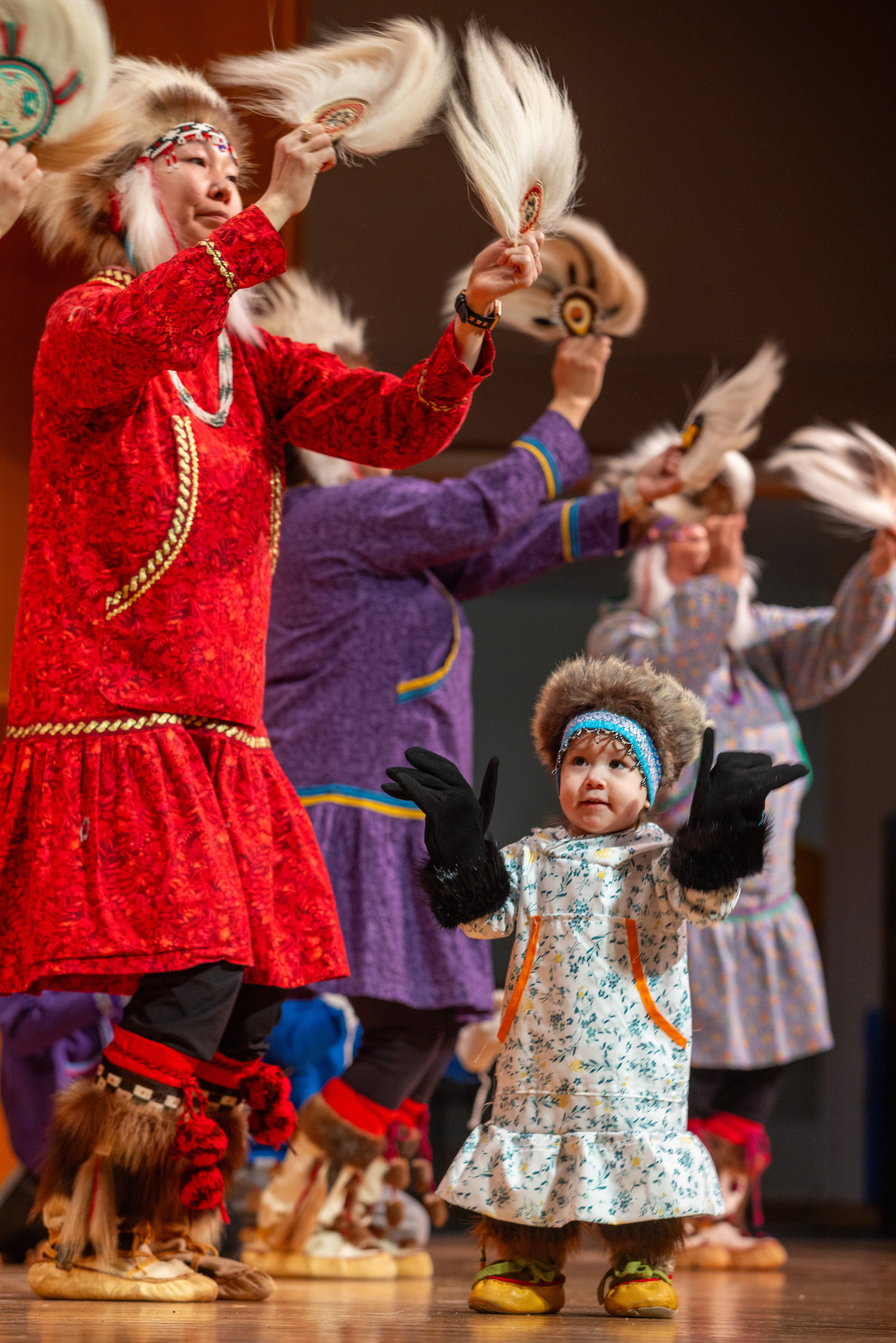 A small child dressed in a kuspuk and regalia dances on a stage in front of a group of Alaska Native dancers.