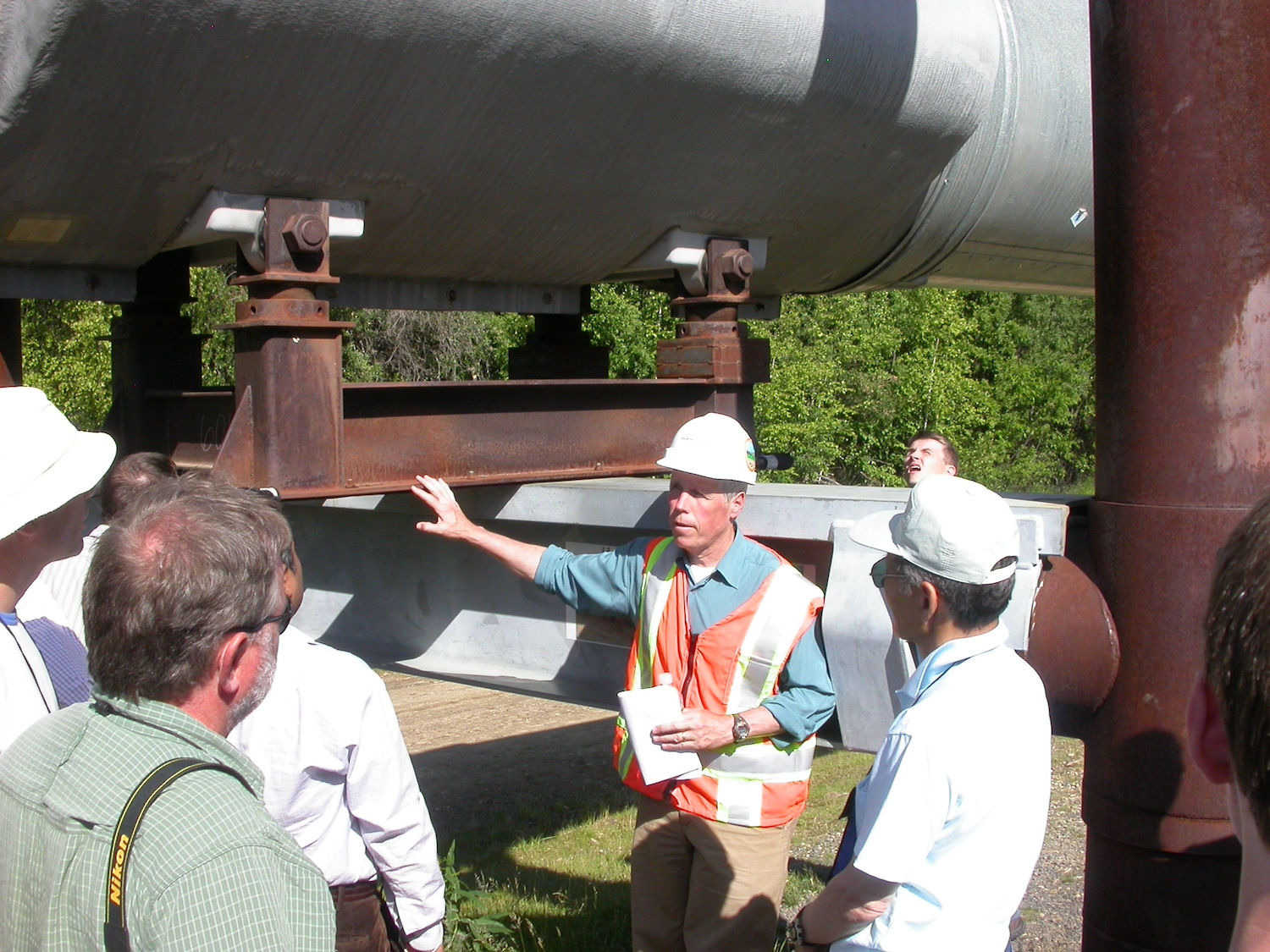 A man in a white hardhat and orange safety vest talks while pointing to a metal structure under a large pipe. Other men stand around him listening.