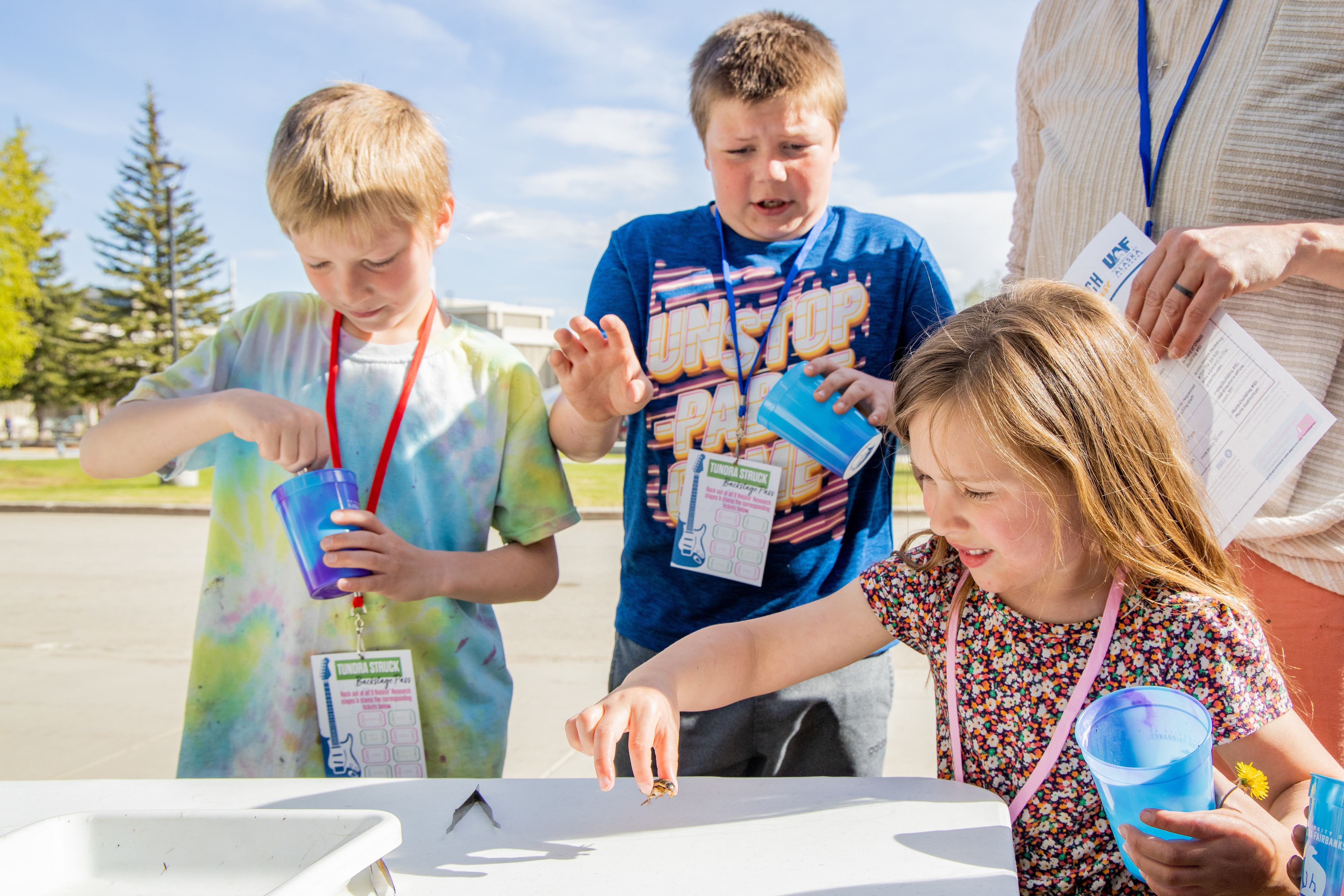 Three children holding cups gathered around a table. One of the children is holding a bug. 