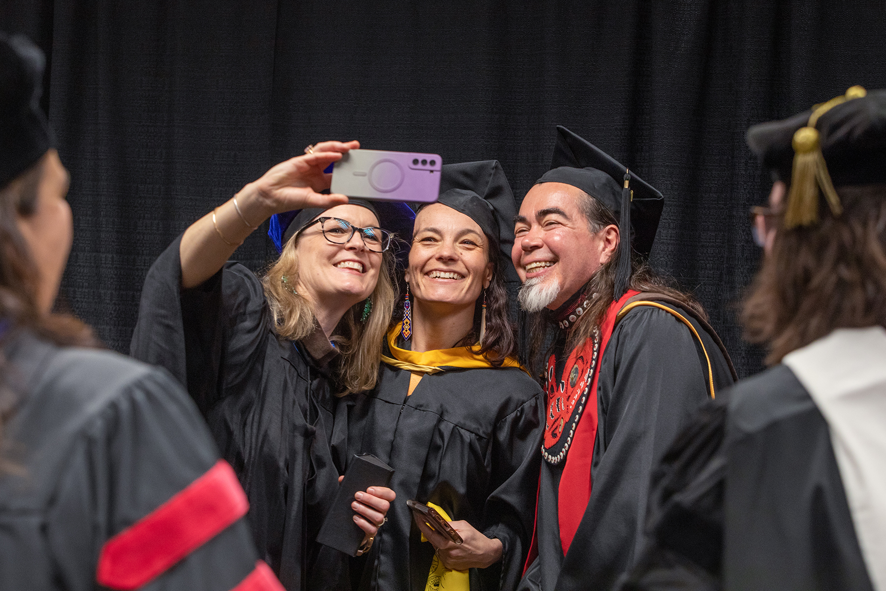 CLA Faculty (from left) Carrie Baker, Professor and Associate Dean, College of Liberal Arts, Dept. of Theatre and Film, Maya Salganek, Director of FRAME and Associate Professor, Film/Video Arts, and Da-ka-xeen Mehner, Professor of Native Art and Department Chair, take a photo before the faculty processional during the University of Alaska Fairbanks 2023 Commencement Ceremony at the Carlson Center Saturday, May 6, 2023. | UAF Photo by Eric Engman